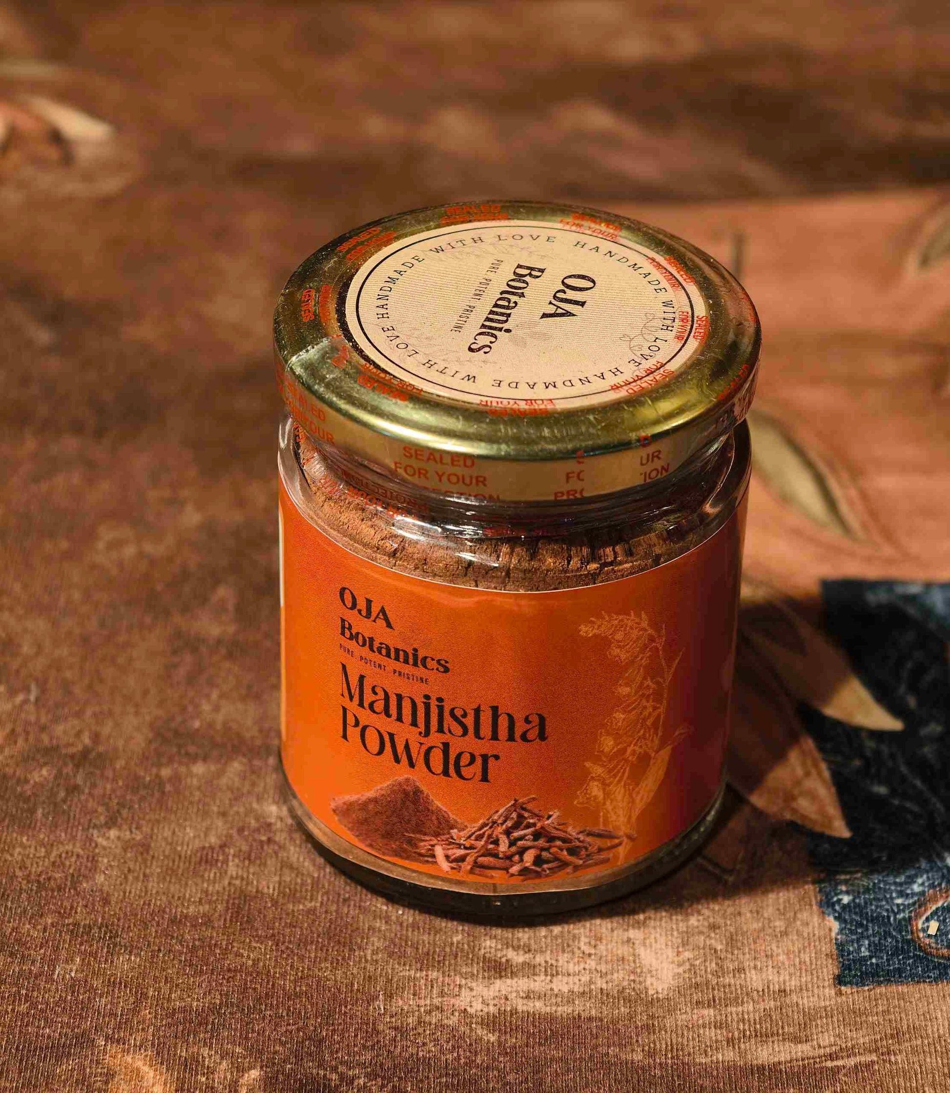 Jar of Oja Botanics Manjistha Powder on a textured fabric background